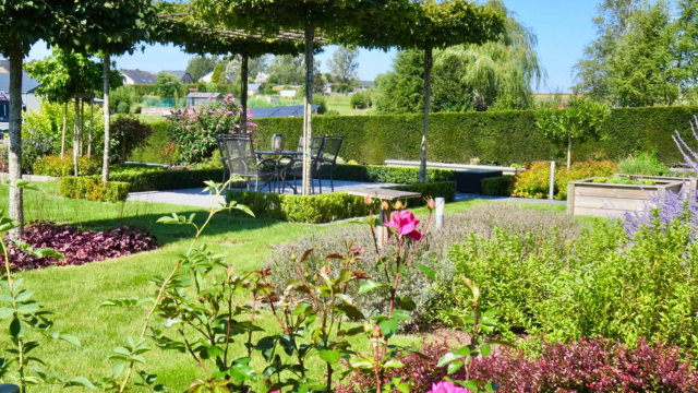 Vue d'un jardin avec des parterres de fleurs colorées, des pelouses et des arbres formant une zone ombragée.